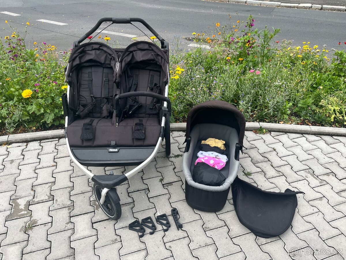 Černý dvoumístný sportovní kočárek Thule vedle korbičky a adaptérů na chodníku